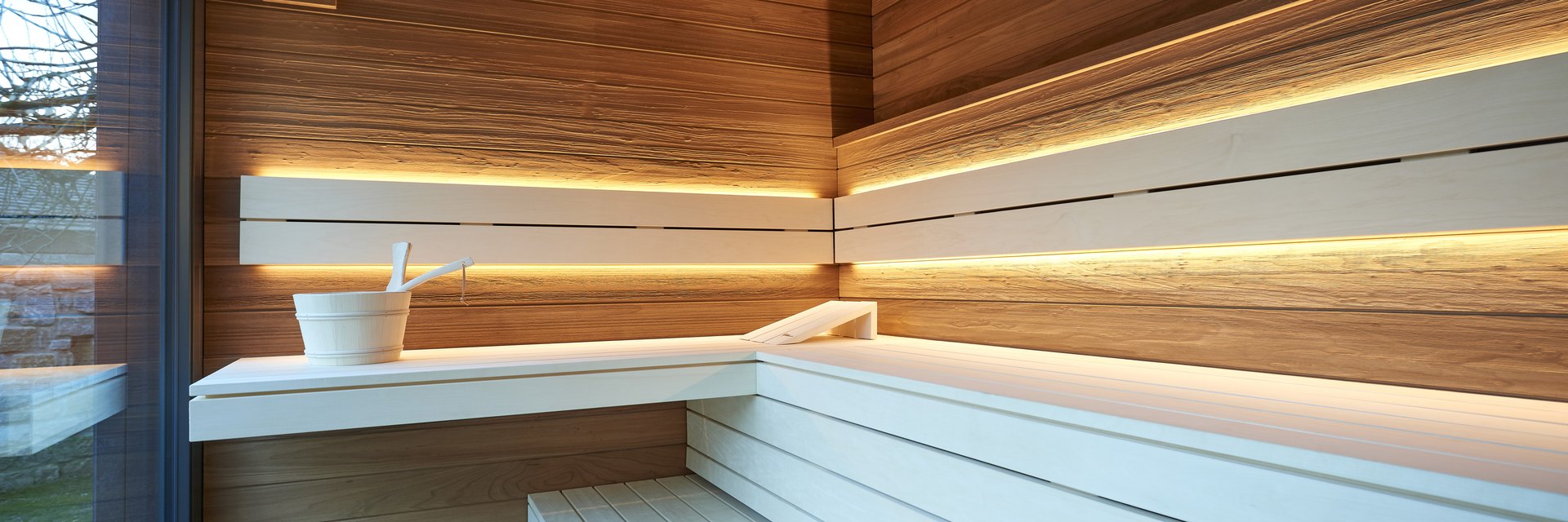 Innenraum einer Designsauna: helle Liegeb&auml;nke vor Zedernholzwand, umlaufendes indirektes Lichtband und Saunak&uuml;bel, daneben die raumhohe Glasfront in den Garten