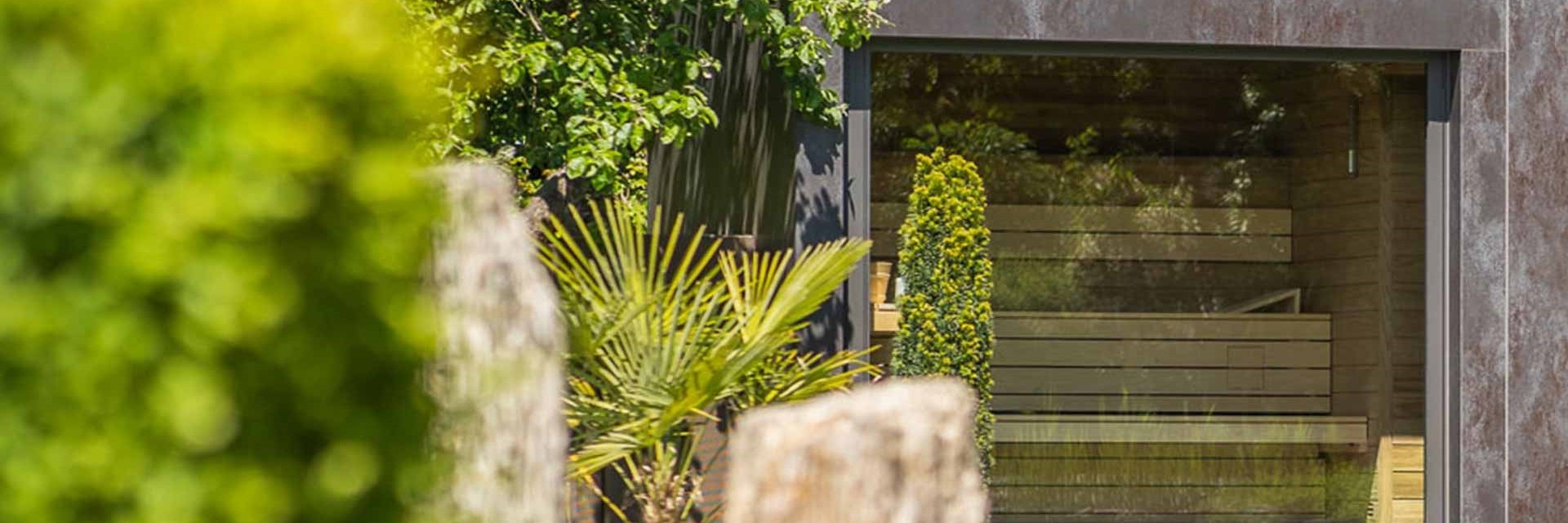 Moderne Gartensauna mit steinfarbener Fassade und gro&szlig;em Panoramafenster, dahinter die Saunab&auml;nke, davor Palmfarn, Buchsformschnitt und Steinstelen