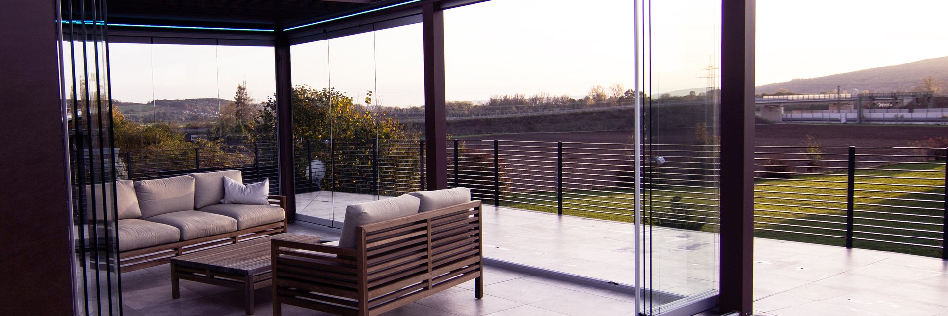 Terrassen&uuml;berdachung mit rahmenlosen Glasschiebeelementen, dahinter eine Loungegruppe aus Holz und der Blick &uuml;ber die Felder in die Abendsonne