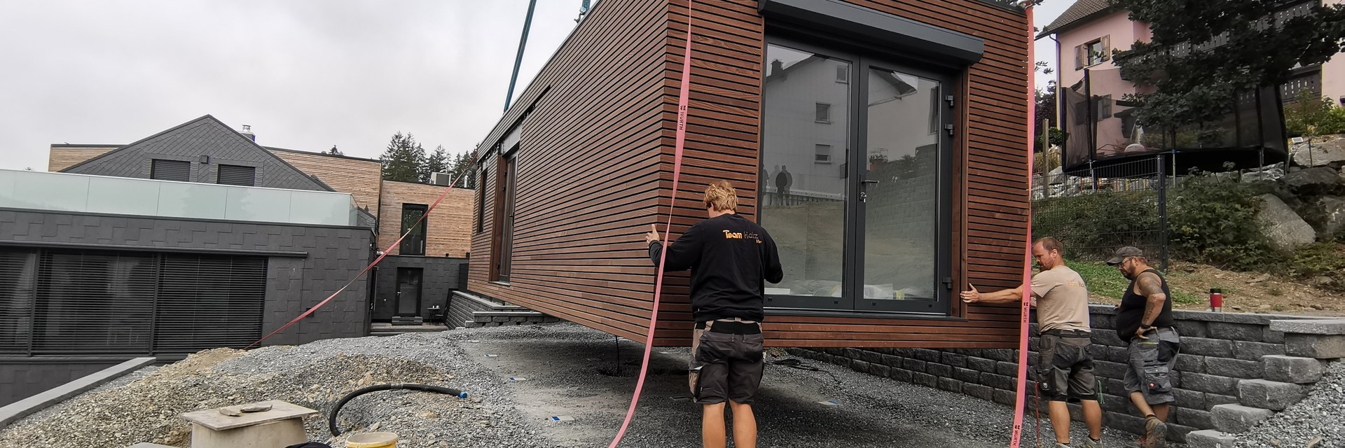 Drei Handwerker setzen ein vorgefertigtes Holzmodul am Kran auf die vorbereitete Natursteinmauer