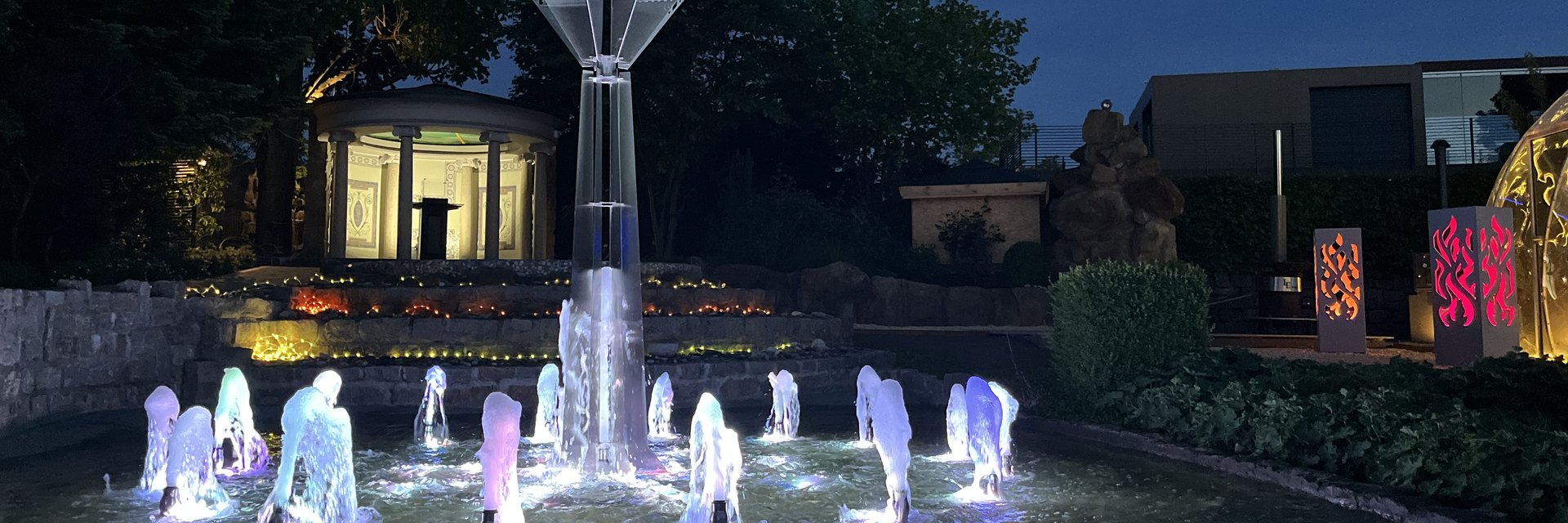 Beleuchtetes Wasserspiel aus Edelstahl bei D&auml;mmerung: farbig angestrahlte Font&auml;nen in einem Becken vor der beleuchteten Treppenanlage des LE Showrooms