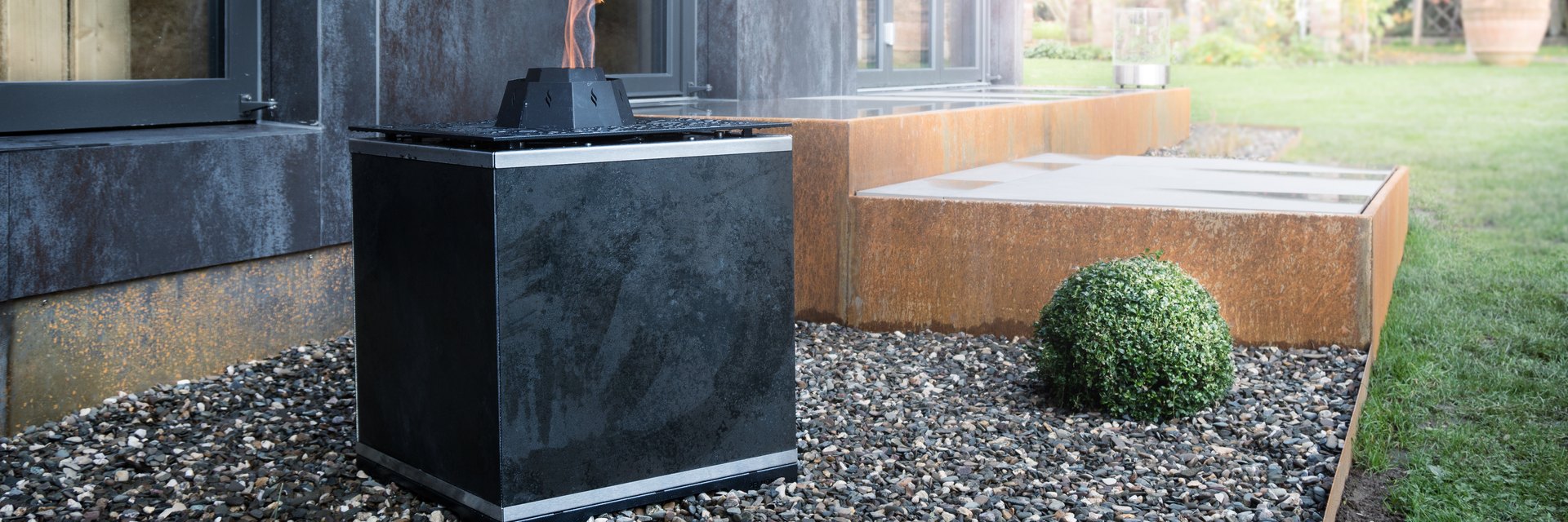 LE FireCube: wuerfelfoermige Feuerstelle mit dunkler Steinverkleidung und offener Flamme im Kiesbeet vor einer Cortenstahl-Terrasse