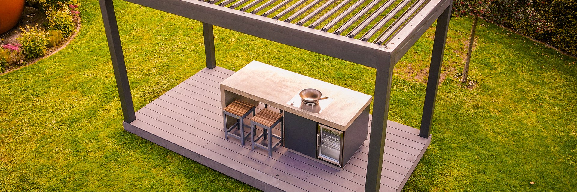 Ausgef&uuml;hrter LE KitchenCube aus erh&ouml;hter Sicht: Pergola mit geschlossenem Lamellendach auf einem Holzdeck in der Rasenfl&auml;che, darunter Kochinsel mit Wok-Brenner, Barhockern und Weink&uuml;hlschrank