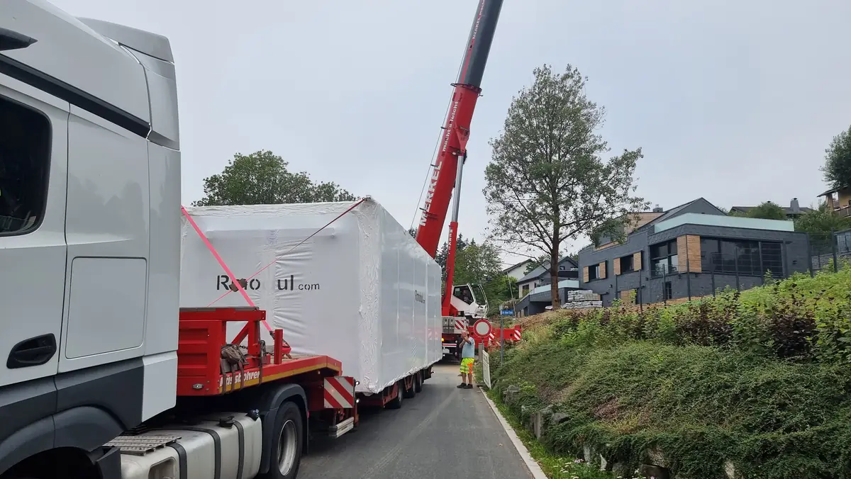 Das in Folie verpackte Wohnmodul auf dem Tieflader in einer Dorfstraße, dahinter der Autokran