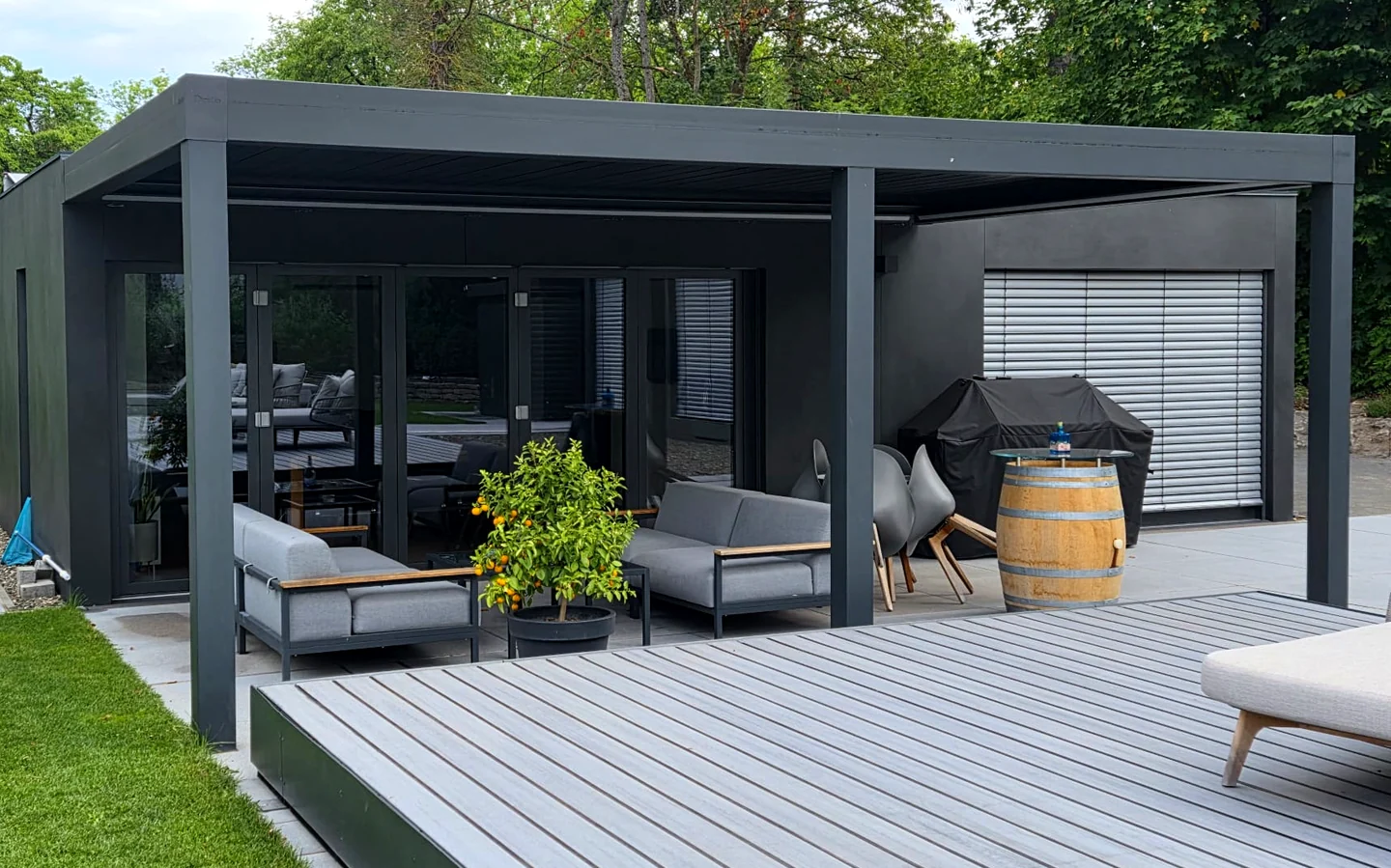 Realisiertes LE Poolhouse: dunkler Baukörper mit raumhoher Glas-Faltwand, Pergola und Lounge auf dem Terrassendeck