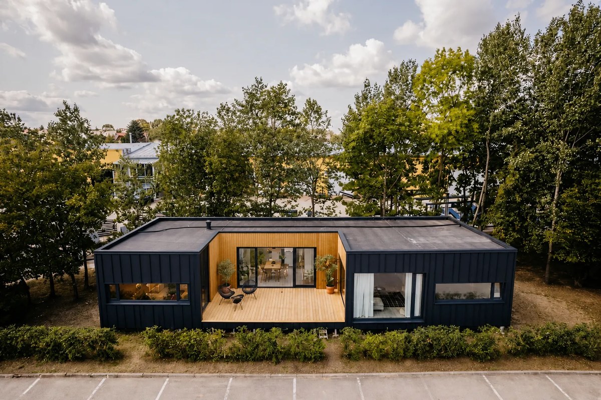 Ausgef&uuml;hrtes LE ModulHaus in der Frontalansicht: anthrazitfarbene Stehfalzfassade, mittig zur&uuml;ckgesetzte Holzterrasse mit raumhoher Verglasung, umgeben von B&auml;umen