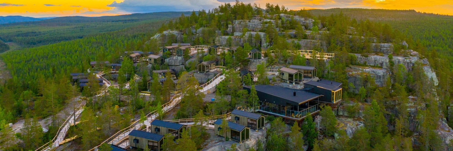 Luftaufnahme einer Siedlung aus vorgefertigten Holzmodulbauten auf einem Felsplateau im Nadelwald, dazwischen Stege und ein gr&ouml;&szlig;eres Gemeinschaftsgeb&auml;ude mit verglaster Terrasse, im Abendlicht