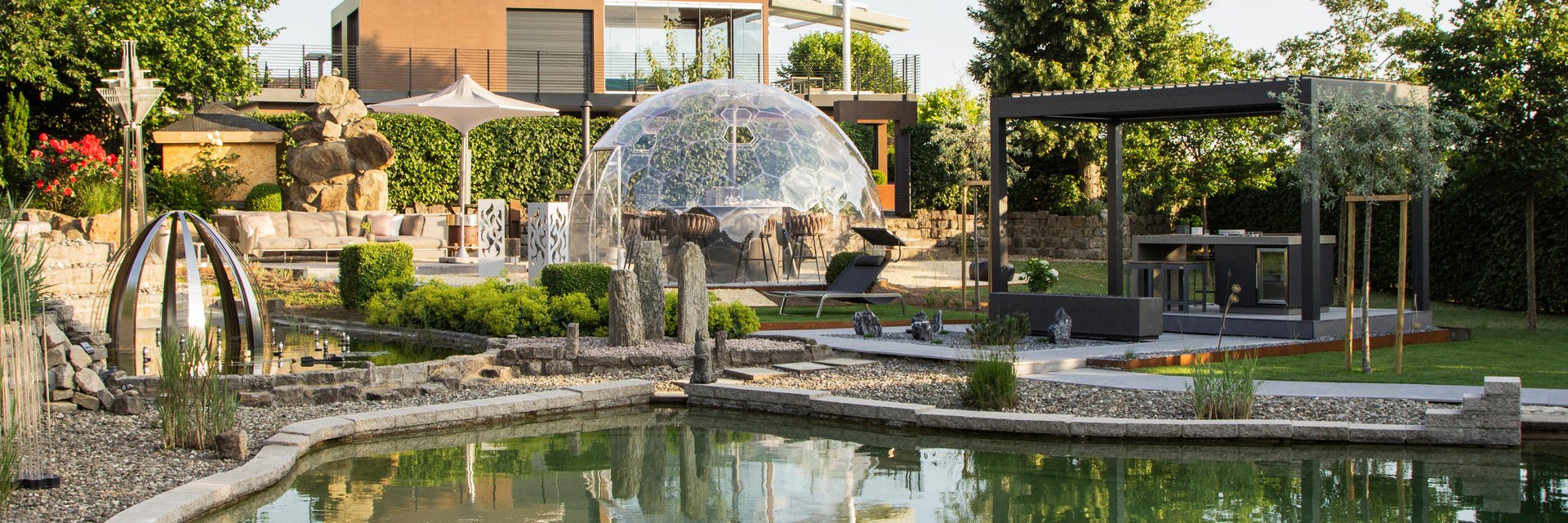 LE Showroom im Abendlicht mit Naturteich, Gartenzimmer unter Glaskuppel, Pergola und Outdoor-K&uuml;che