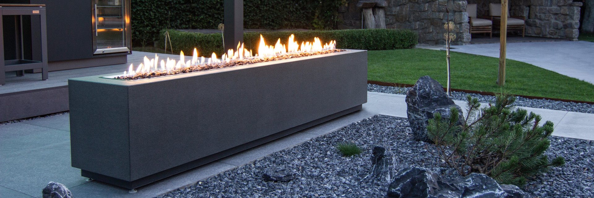 LE FireBox: langer Feuertisch aus Beton mit durchgehender Flammenreihe im Kiesbeet des LE Showrooms in der Daemmerung