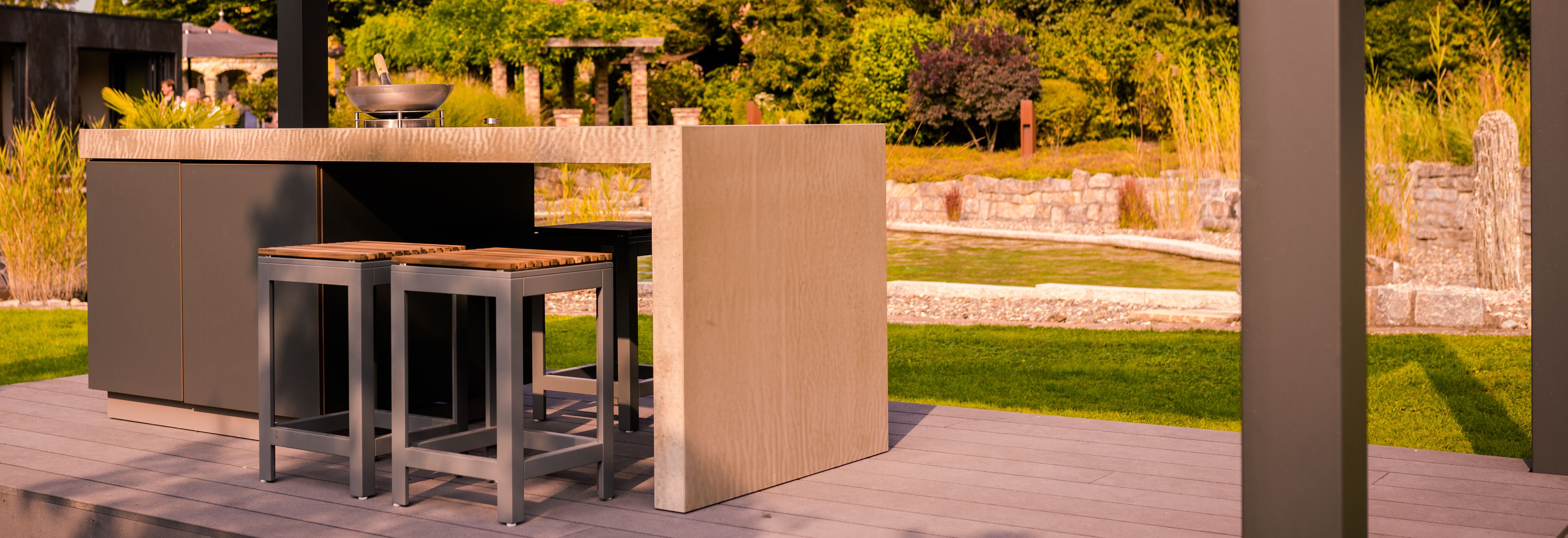 Au&szlig;enshowroom Ebensfeld: Outdoor-K&uuml;che mit Feuerschale und Barhockern auf Holzdeck unter dem Lamellendach, dahinter Pergolagang, Trockenmauern, Gr&auml;ser und ein Wasserbecken