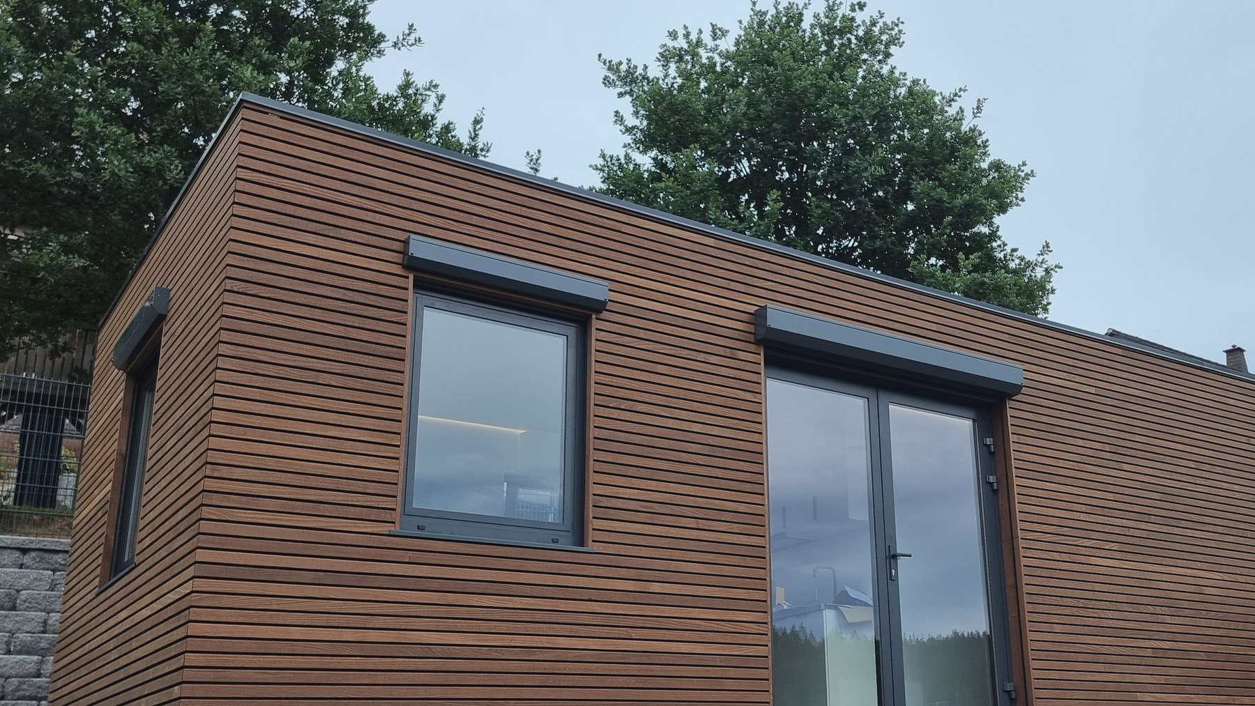 Ausgef&uuml;hrtes LE MicroHouse am Montagetag: Modulbau mit horizontaler Thermoholz-Fassade, Eckausbildung, Fenster und raumhoher Glast&uuml;r mit Raffstore-K&auml;sten, dahinter St&uuml;tzmauer und Baumbestand