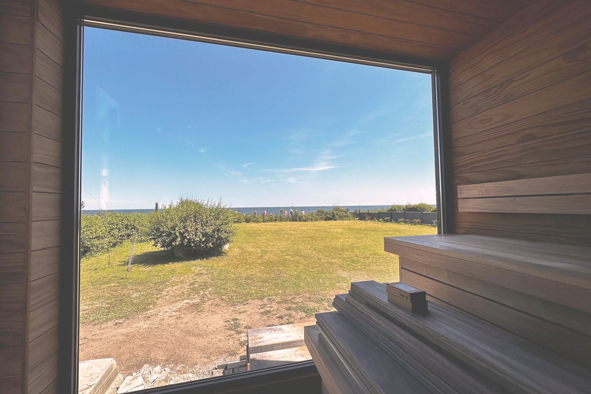 Ausgef&uuml;hrte LE Gartensauna von innen: B&auml;nke und Wandverkleidung aus Holz, raumhohes Panoramafenster mit Blick &uuml;ber die Wiese zur K&uuml;ste