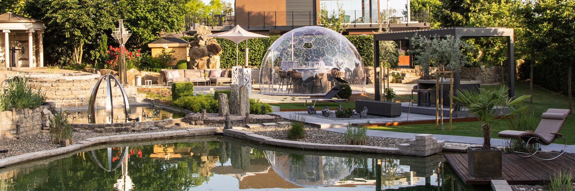 LE Showroom im Abendlicht: Schwimmteich, gl&auml;sernes Kuppelzelt, Lamellenpergola mit Loungem&ouml;beln und Sonnensegel im Schaugarten