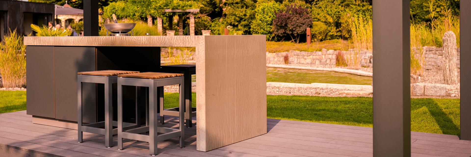 Au&szlig;enshowroom Ebensfeld: Outdoor-K&uuml;che mit Feuerschale und Barhockern auf Holzdeck unter dem Lamellendach, dahinter Pergolagang, Trockenmauern, Gr&auml;ser und ein Wasserbecken