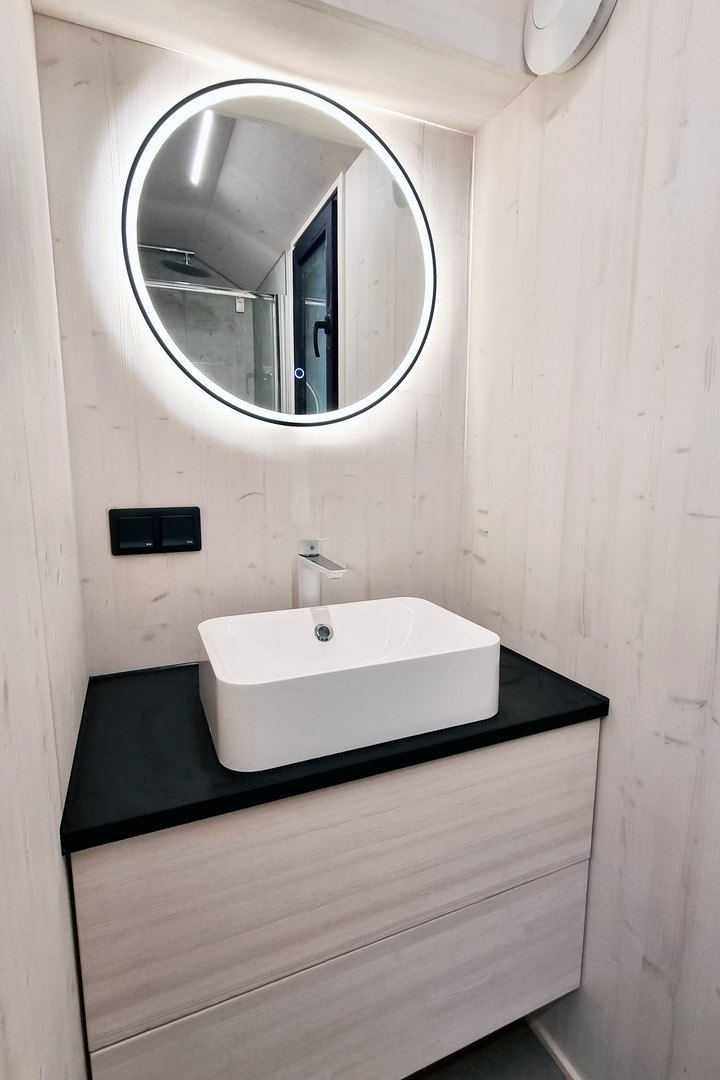 Bad eines ausgef&uuml;hrten LE TinyHouse: wei&szlig;es Aufsatzbecken auf dunkler Platte, dar&uuml;ber ein runder, hinterleuchteter Spiegel