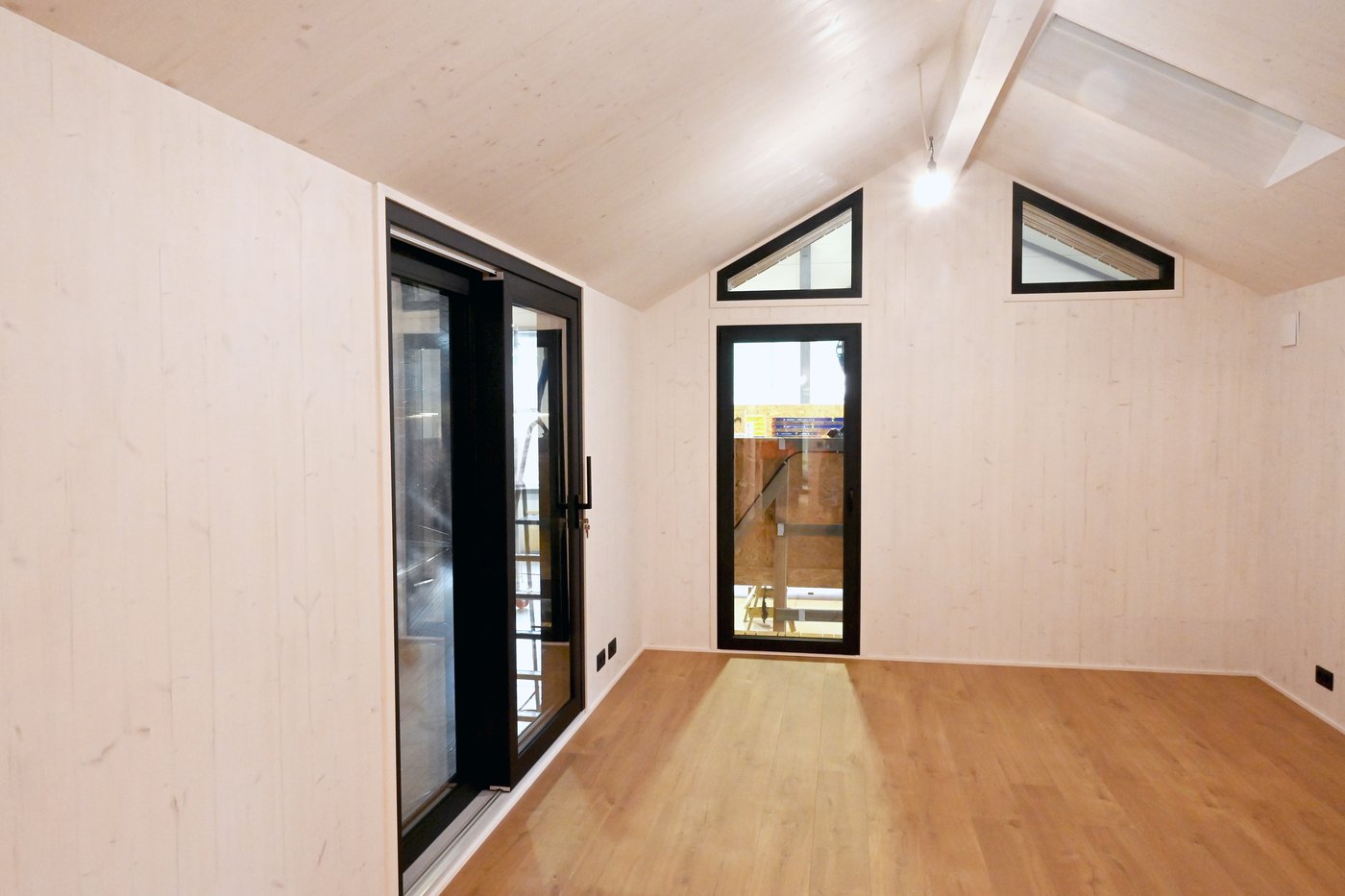 Wohnbereich eines ausgef&uuml;hrten LE TinyHouse: Giebelwand mit Eingangst&uuml;r, zwei dreieckigen Giebelfenstern und raumhoher Verglasung zur L&auml;ngsseite