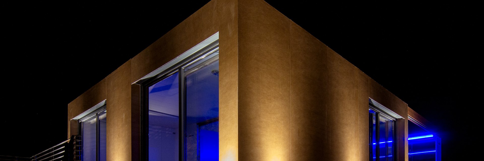 N&auml;chtliche Lichtplanung: Geb&auml;udegiebel mit warmen Bodenstrahlern an der Fassade und blauen Akzentleuchten entlang der Verglasung