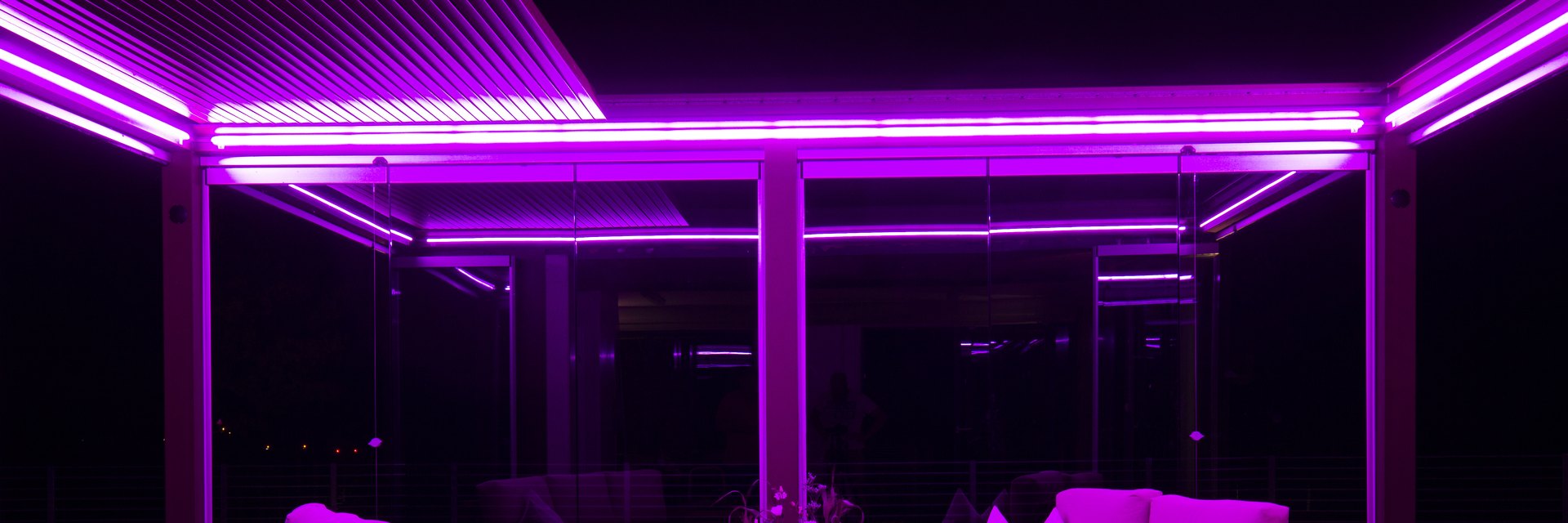 &Uuml;berdachter Au&szlig;enbereich der LE Lounge bei Nacht: magenta leuchtende LED-Linien in der Lamellendecke &uuml;ber Loungem&ouml;beln