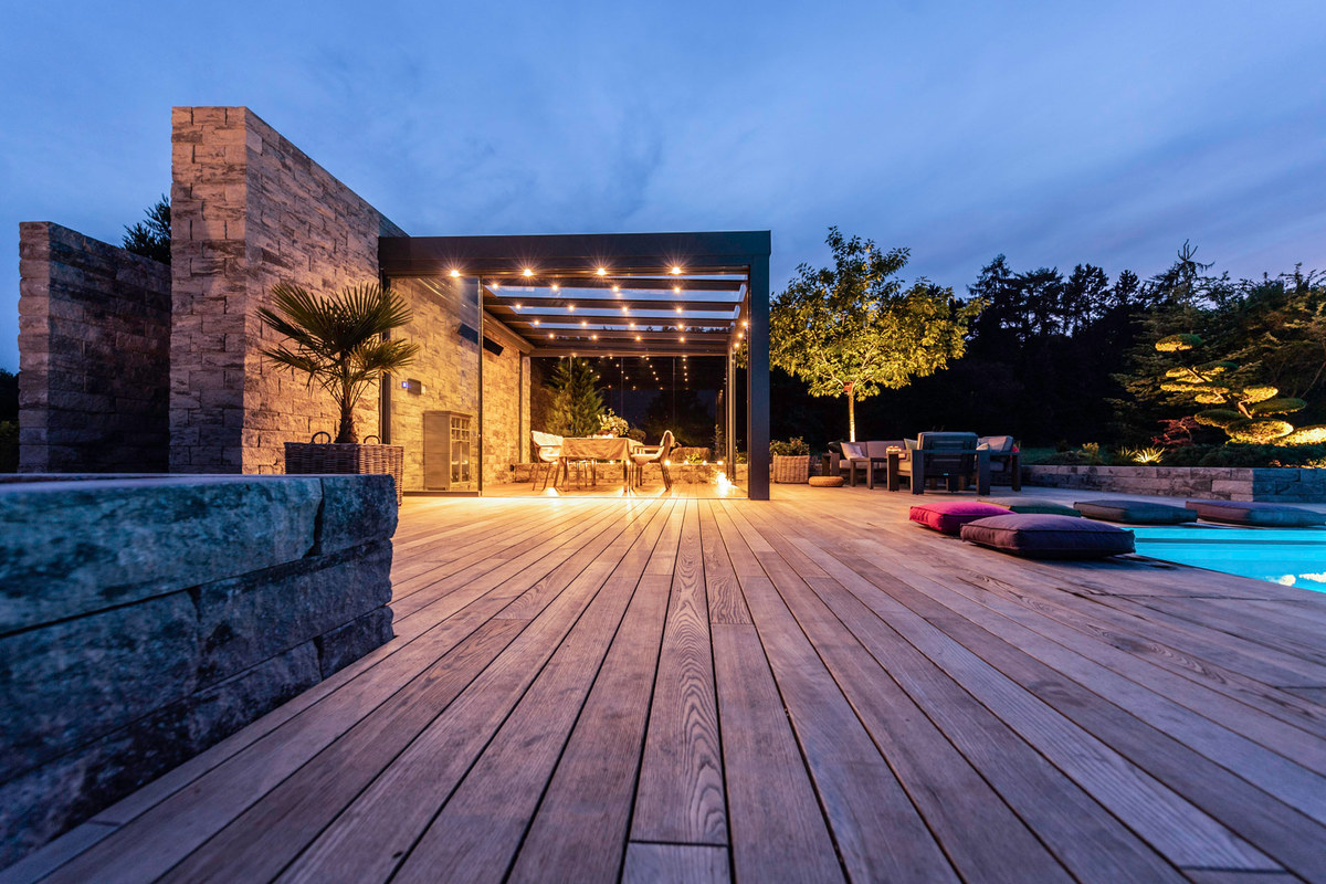Terrassendach mit beleuchtetem Glasdach &uuml;ber einer Holzterrasse am Pool in der Abendd&auml;mmerung