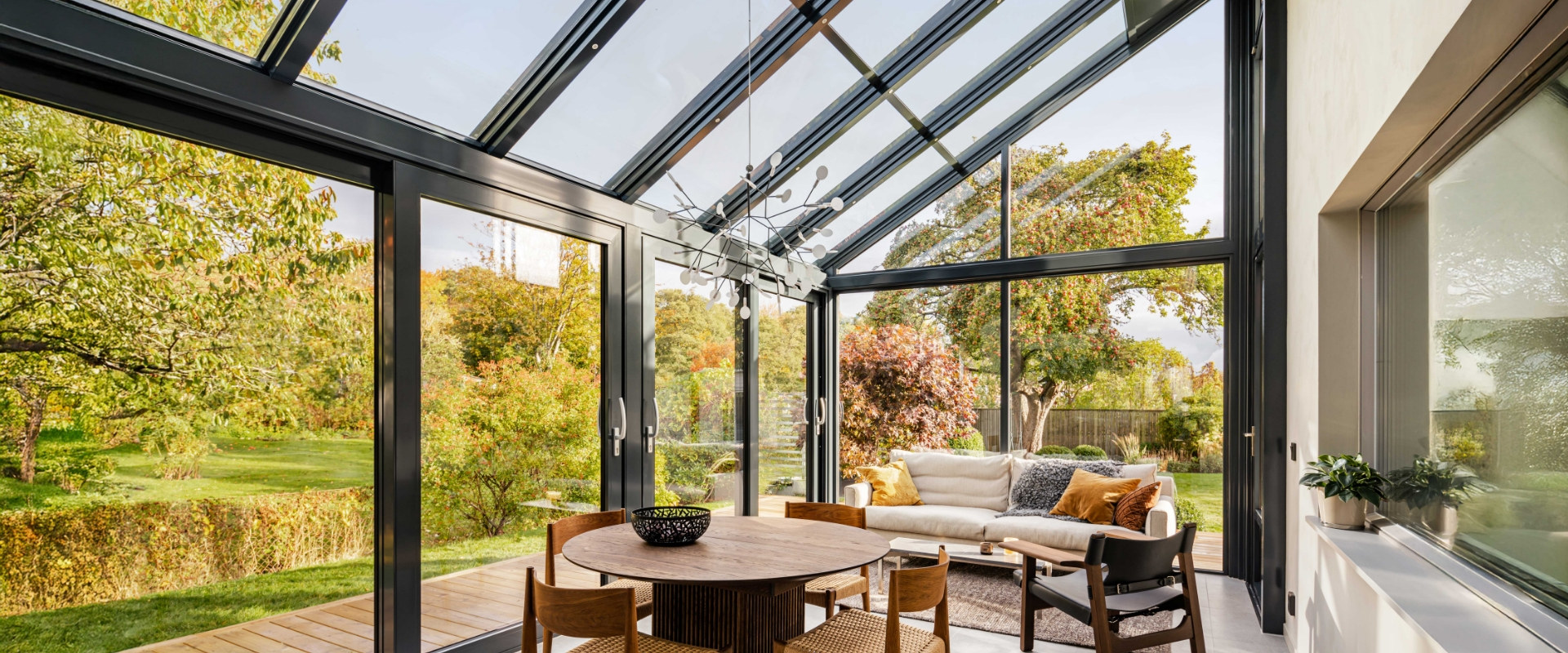 Wintergarten mit Glasdach und schwarzen Profilen, innen Esstisch und Sofa, Blick in einen herbstlichen Garten