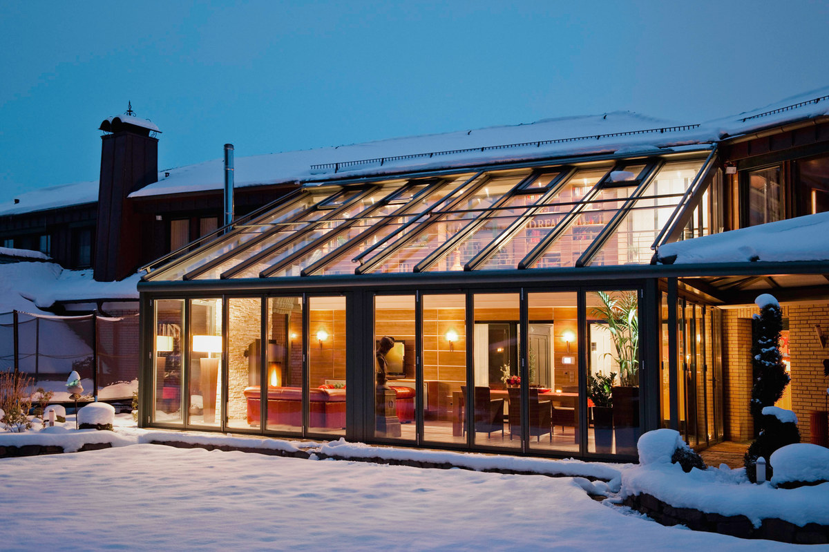 Beleuchteter Wintergarten mit Glasdach vor einem Wohnhaus am Abend, davor liegt Schnee